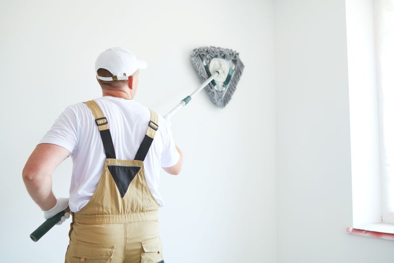 A man cleaning a white wall with a large duster from a distance, wearing beige overalls.