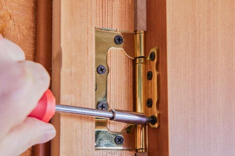 A hand using a screwdriver to tighten screws on a brass door hinge.