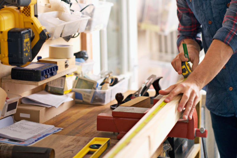 A person measuring a wooden plank with a tape measure in a workshop, surrounded by tools.