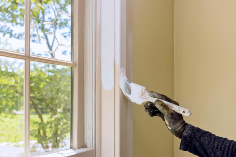 A person painting a window trim with a brush, wearing gloves and a long-sleeved shirt.