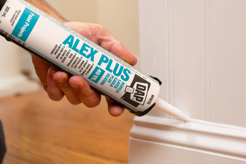 A person applying DAP ALEX PLUS caulk to a white baseboard in a room.
