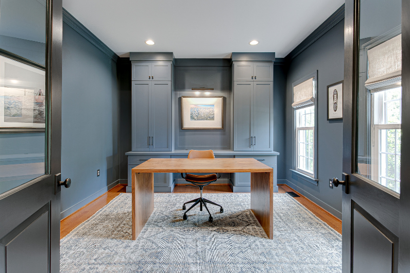 A modern home office with navy blue walls, trim, built-in shelving, wooden desk, and swivel chair, lit by overhead lights.