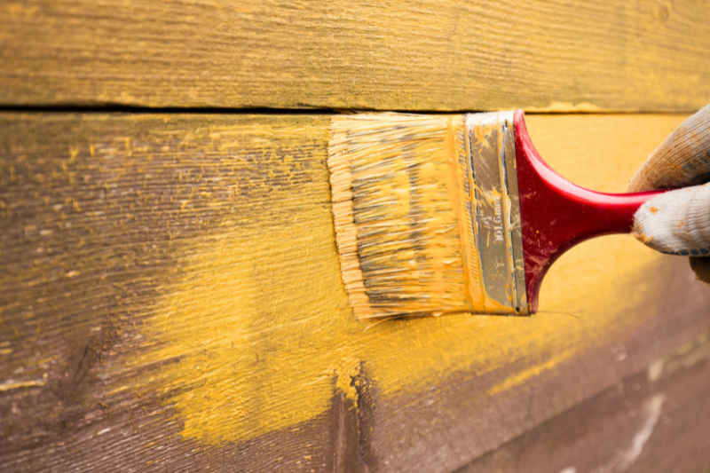 Person using a paint brush to apply bright yellow paint onto wood planks.