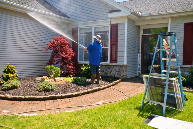 Man using hose to wash siding of house. Ladder to the side, with window screens propped up.