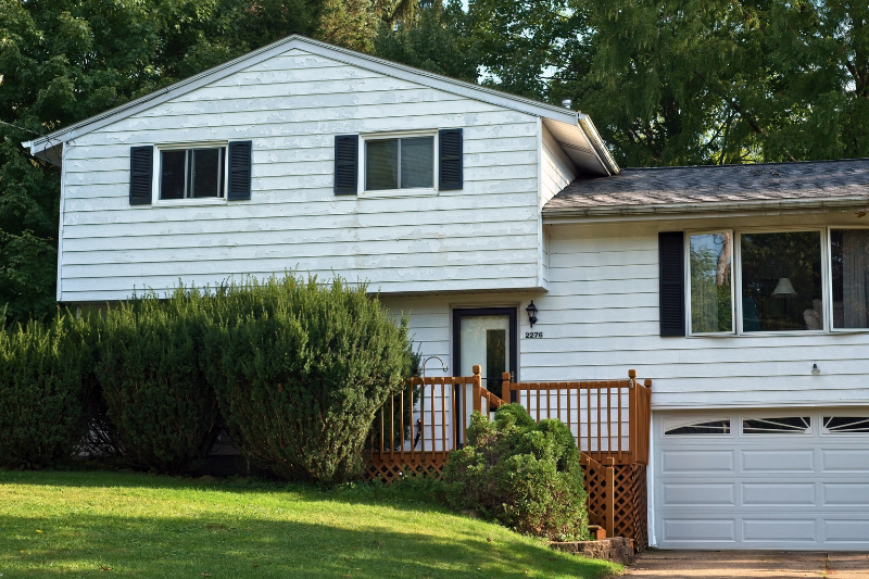 Front of an older home, paint is peeling from the white siding. Overgrown shrubs hide a wooden porch.