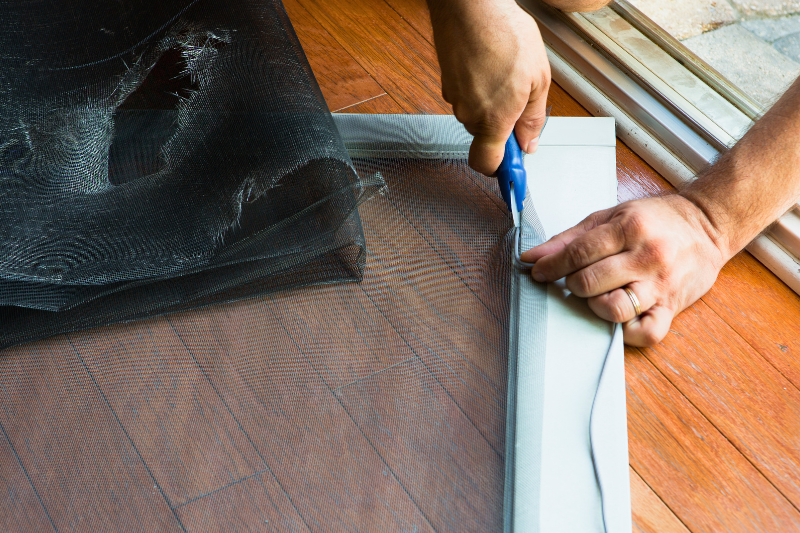 A person trimming excess mesh from a window screen frame. An old screen with a large hole is folded beside.