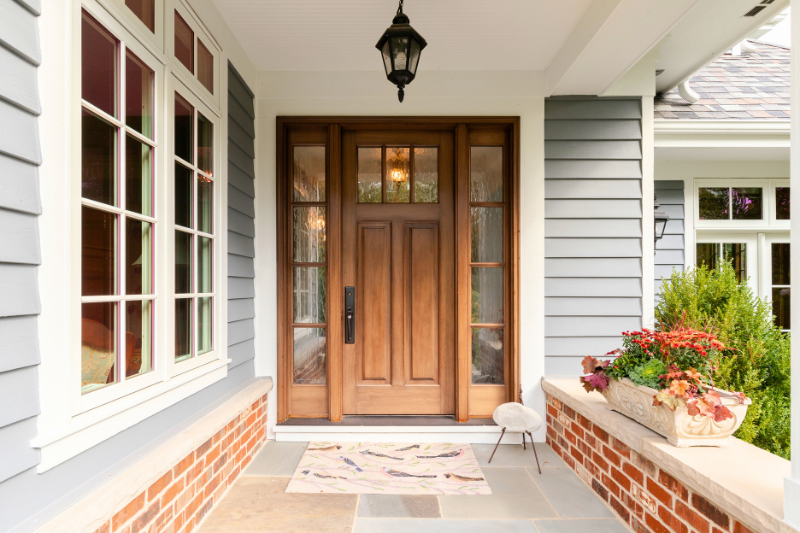 The welcoming front porch of a grey house with a wooden door, potted plants, and red brick accents. A large window with traditional white grilles to the side.