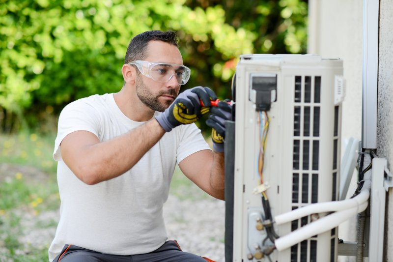 A technician in safety glasses and gloves works on an outdoor air conditioning unit.