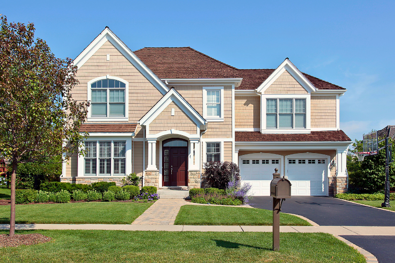 A two-story home with a tan facade, stone accents, and a brown roof, surrounded by green grass and trees. A rich black asphalt driveway leads to a two-car garage.