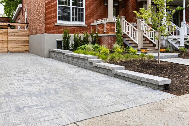 A red brick house with a gray stone pathway, steps, and landscaped plants next to a grey brick herringbone-patterned driveway.