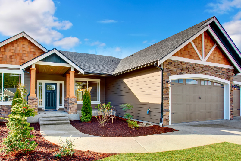 Modern yet rustic home featuring a stone and wood exterior, a front porch, and a garage, surrounded by landscaped shrubs. A curved poured concrete walkway extends from the driveway to the front door.